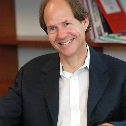 A man is sitting and smiling in an office, wearing a dark blazer over a white shirt; shelves with books and folders are visible in the background.