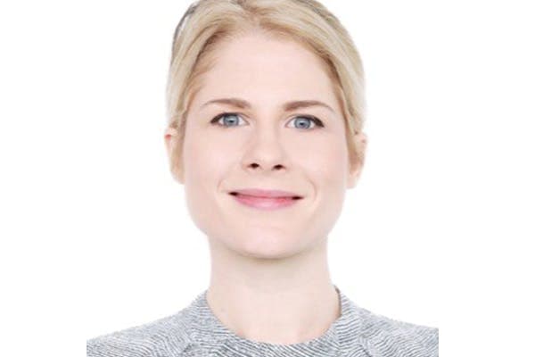 A person with light hair and light blue eyes smiling directly at the camera, wearing a gray patterned top, against a plain white background.