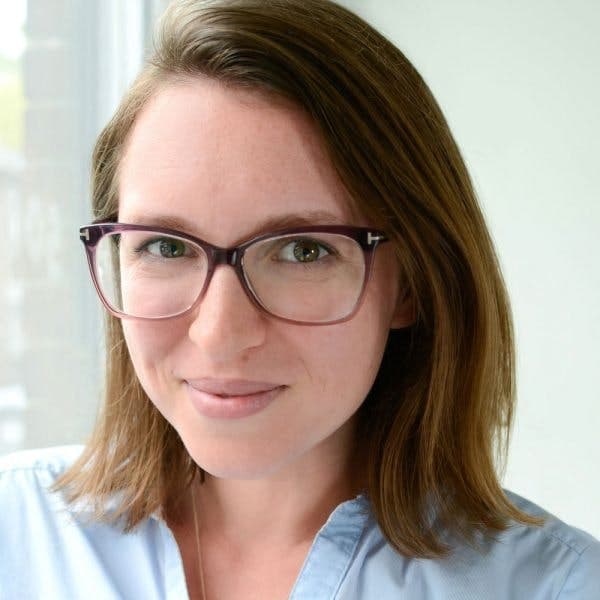 A person with shoulder-length brown hair, glasses, and a light blue shirt, smiling softly, standing inside near a window with a blurred background.