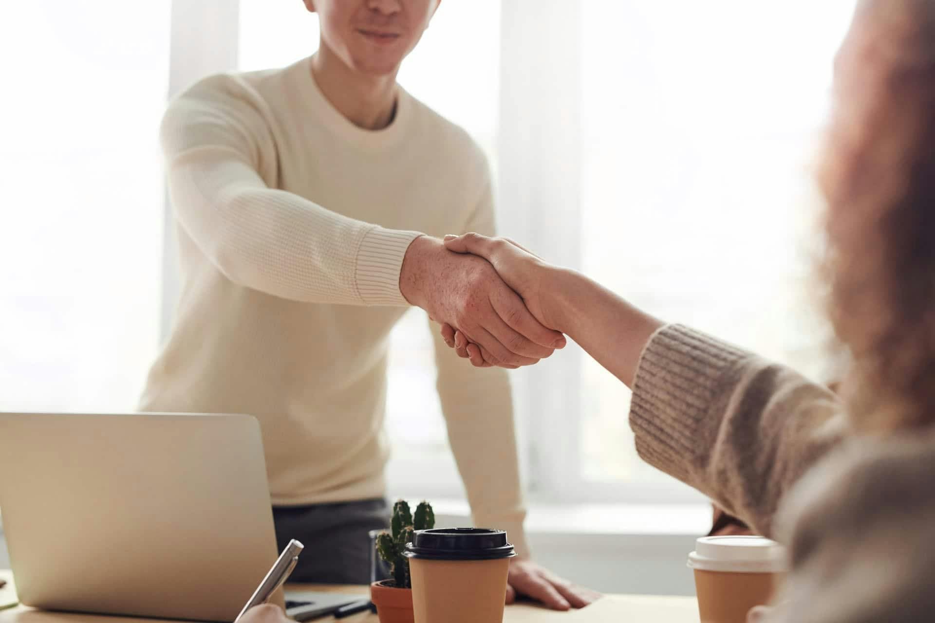 Two people are shaking hands across a table; a laptop, coffee cup, and smartphone are on the table, with a bright window in the background.
