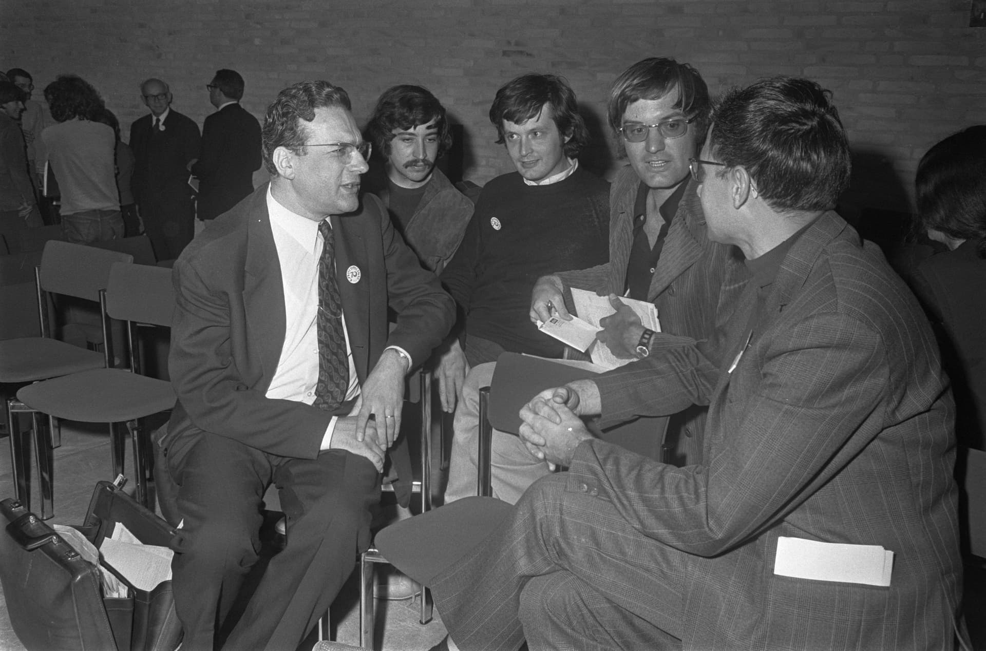 Three men stand closely together, engaging in conversation; one wears glasses and a suit, another has long hair and a mustache, and the third wears a casual shirt with a pin. Background: plain brick wall.