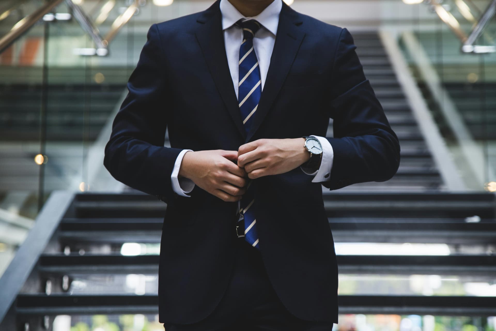 A person adjusts their dark suit jacket while standing on modern indoor stairs, wearing a striped tie and a wristwatch in a professional environment.