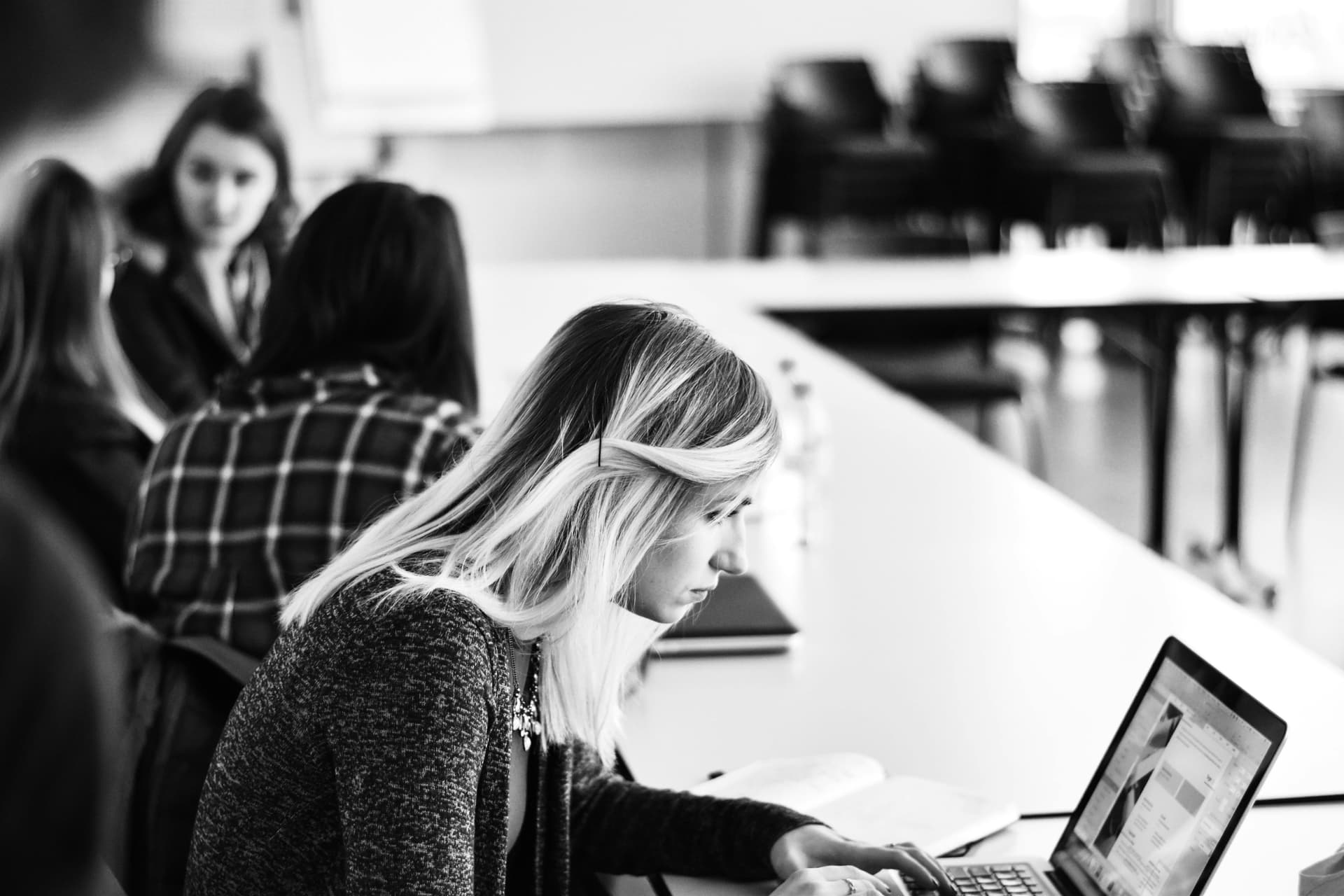 A black-and-white image of a woman working on a laptop at a conference table, with other people conversing in the background classroom environment.