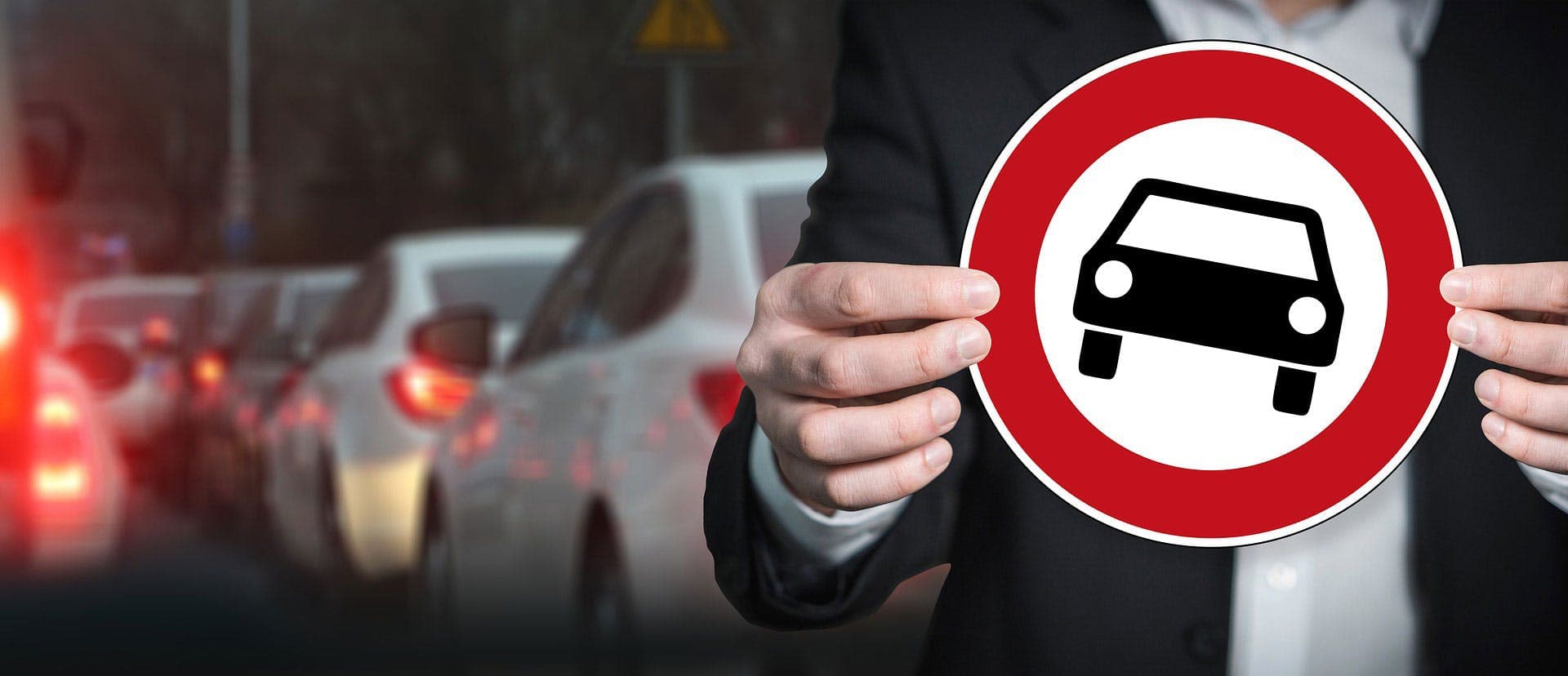 A person holds a "No Entry for Vehicles" sign, standing against a backdrop of stationary cars in traffic on a dimly lit street.