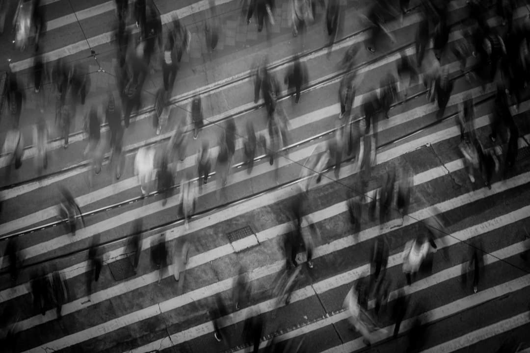 People are crossing a street in a busy urban area, captured with motion blur, depicting fast-paced movement. The crosswalk's striped lines are visible amidst the blurred figures.