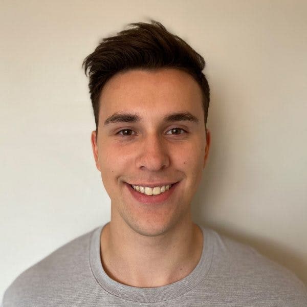 A man is smiling at the camera, wearing a gray shirt, against a plain light-colored background.