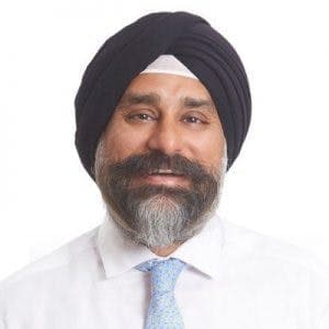 A man with a turban and beard is smiling, wearing a white shirt and blue patterned tie against a plain white background.