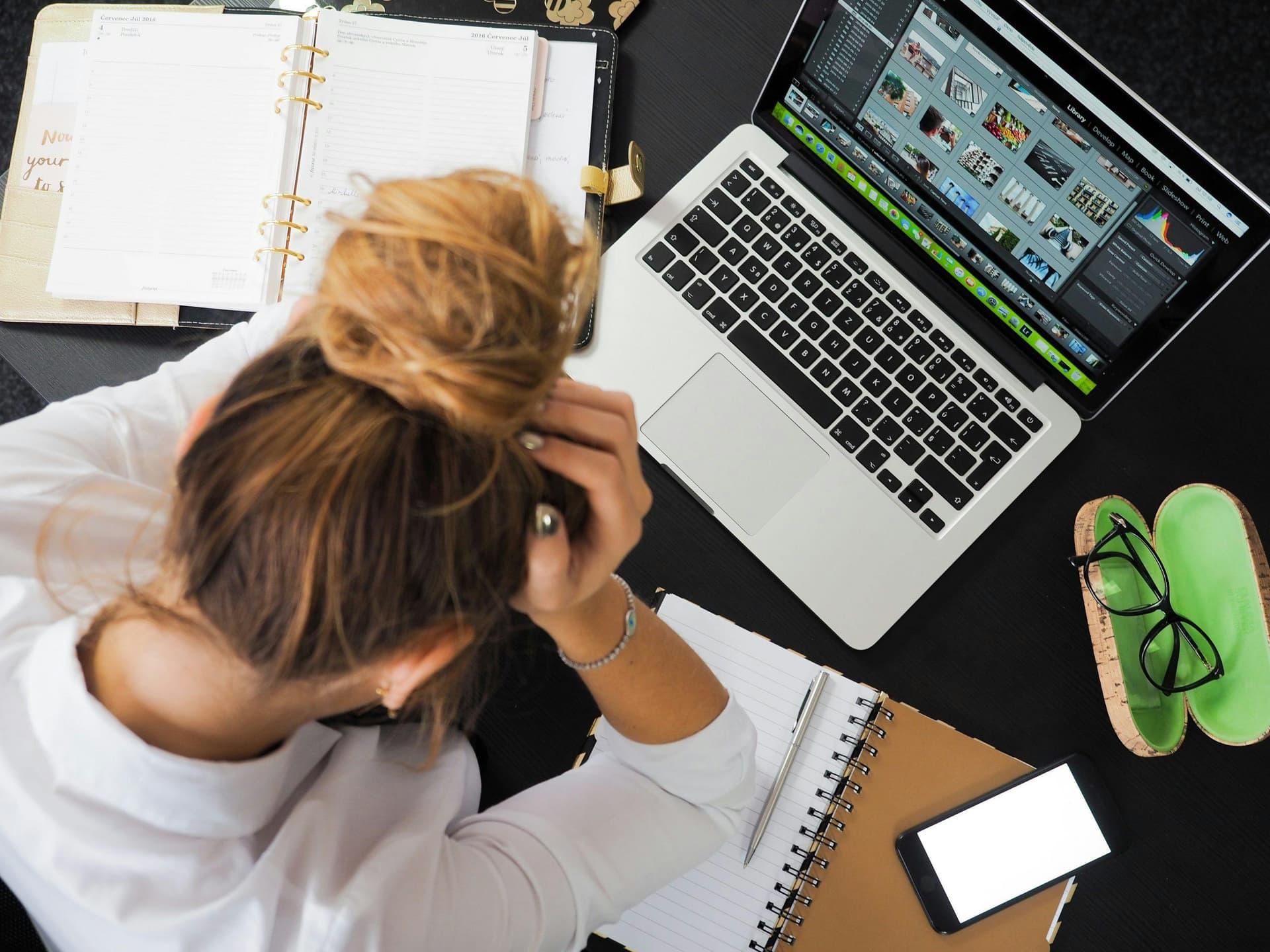 Woman with a messy bun is sitting at a desk with her head in her hands; a laptop showing image thumbnails, notebooks, a notepad, a smartphone, and eyeglasses case surround her.