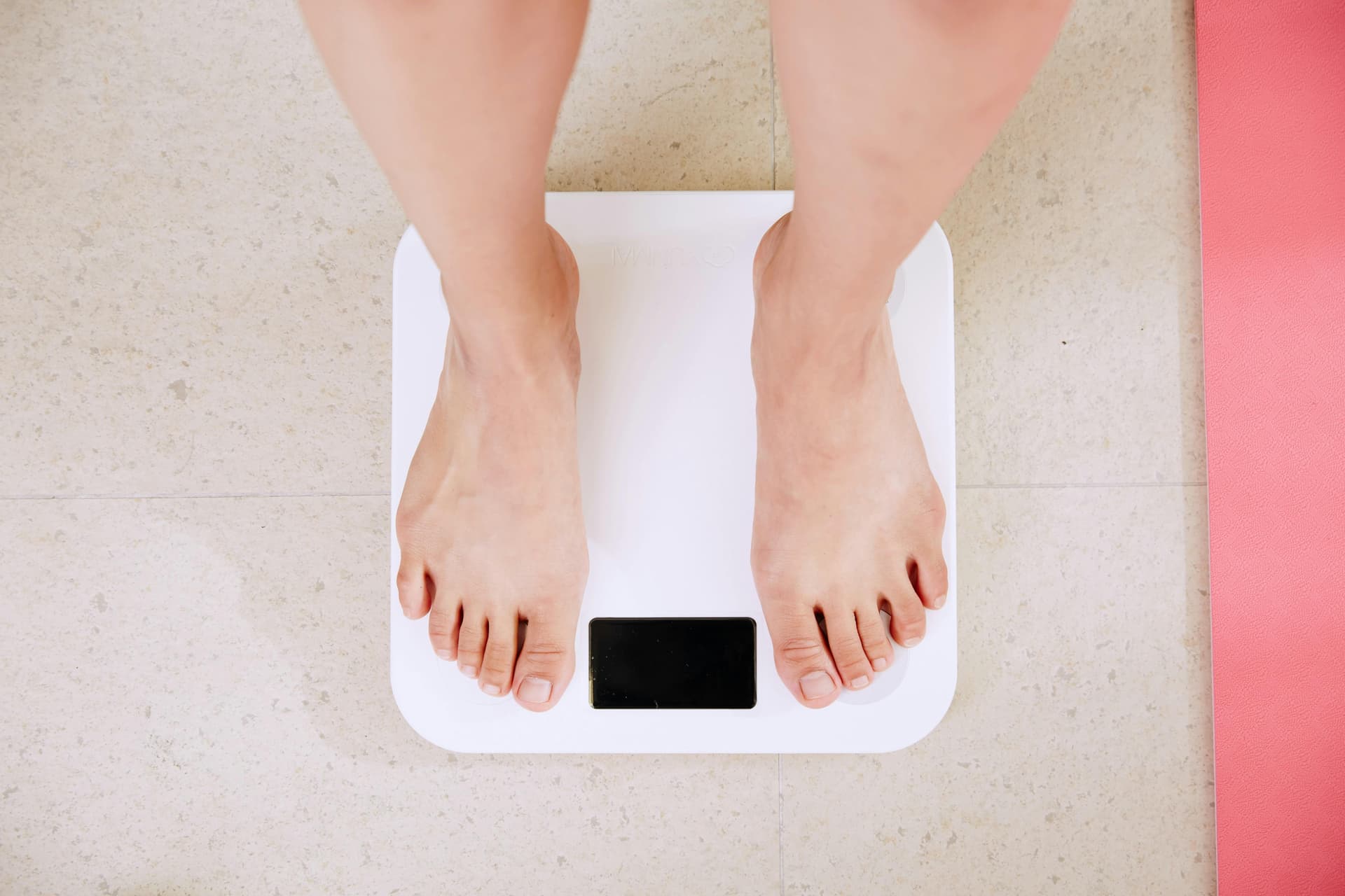 Feet standing on a white digital scale, with a pink yoga mat nearby, on a tiled floor.