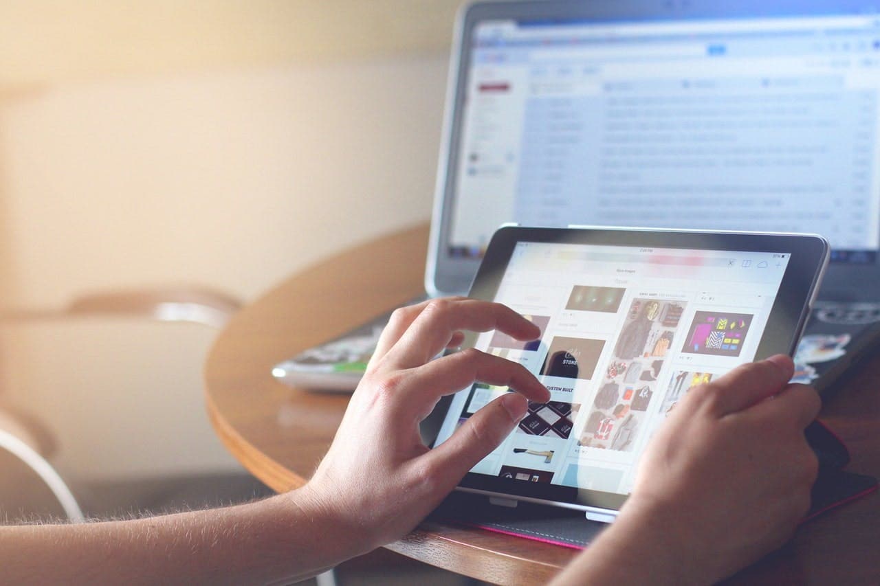 Person using a tablet to browse online images, with a laptop open in the background displaying an email inbox on the screen.