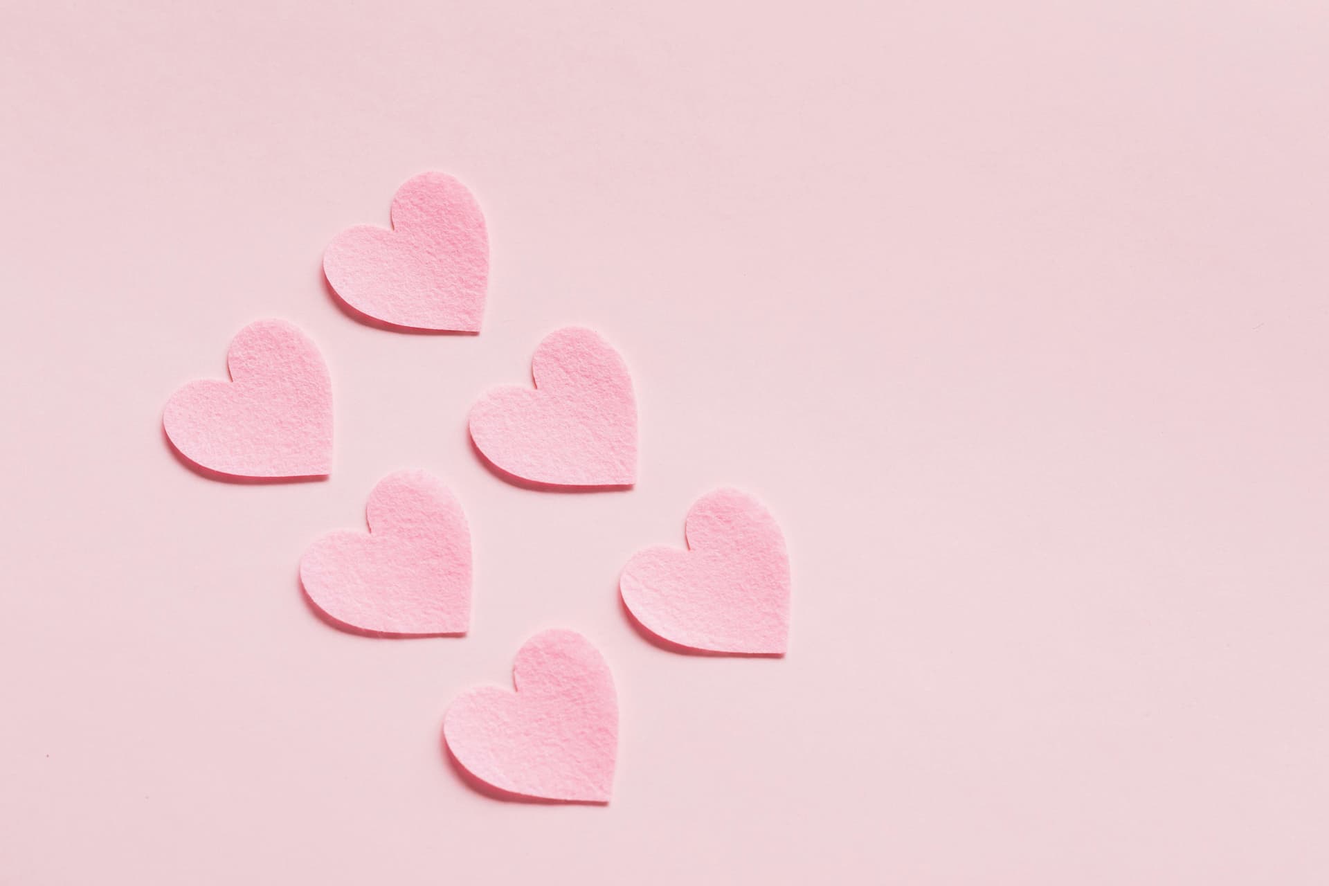 Six pink felt hearts lie scattered on a smooth, light-pink background, creating a simple yet charming design.