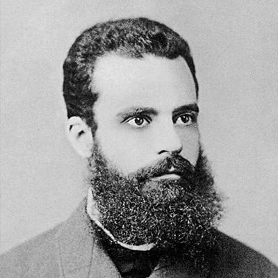 A bearded man with short, wavy hair and a serious expression poses for a black-and-white portrait; he wears a high-collared suit jacket and stands against a plain background.