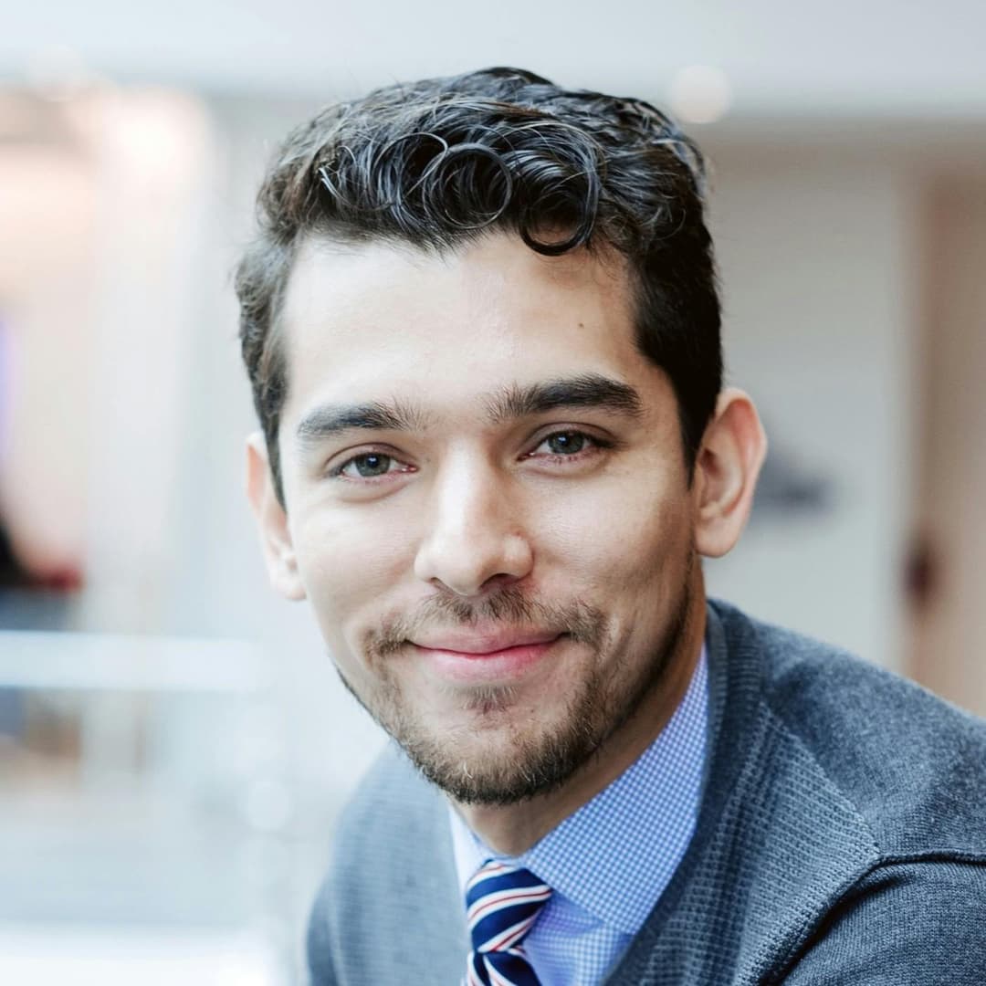 A man with dark, curly hair and a beard smiles, wearing a blue dress shirt, striped tie, and grey sweater, in a bright indoor setting with blurred background.