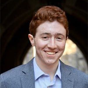 A young man with red hair and a blue shirt under a grey suit jacket is smiling, standing in front of a dark archway with blurred sunlight in the background.