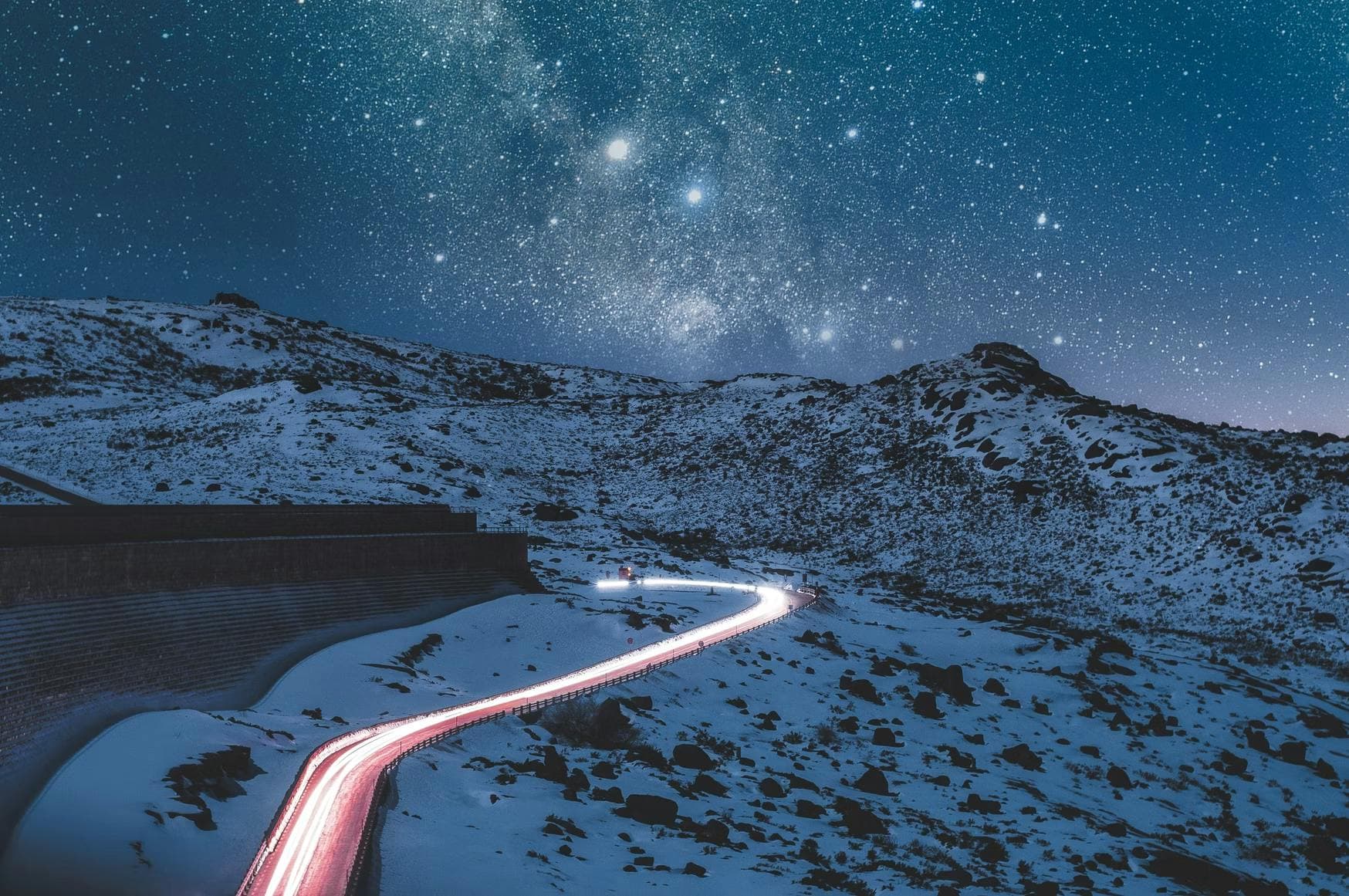 A winding road, illuminated by car lights, cuts through a snowy, mountainous landscape under a vast, star-filled night sky.
