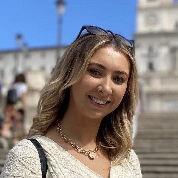 A smiling woman wearing sunglasses on her head and a necklace stands in front of a staircase with blurred historical buildings and people in the background.