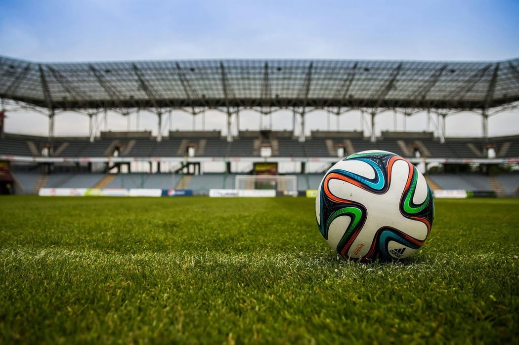 A colorful soccer ball rests on a grass field, with empty stadium seating in the background. 