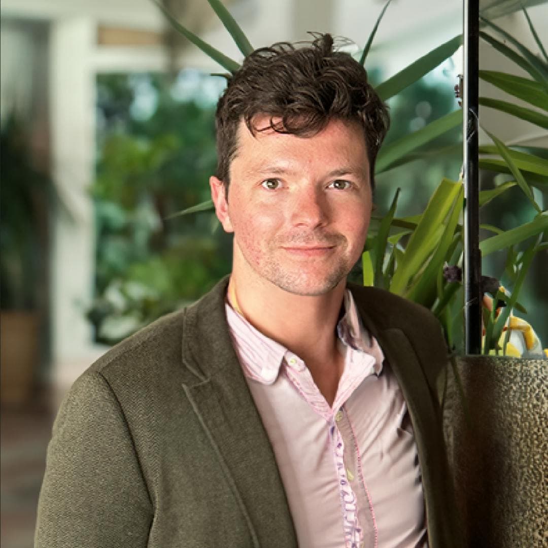 A man with curly hair, wearing a gray suit over a pink button up shirt looking directly ahead.