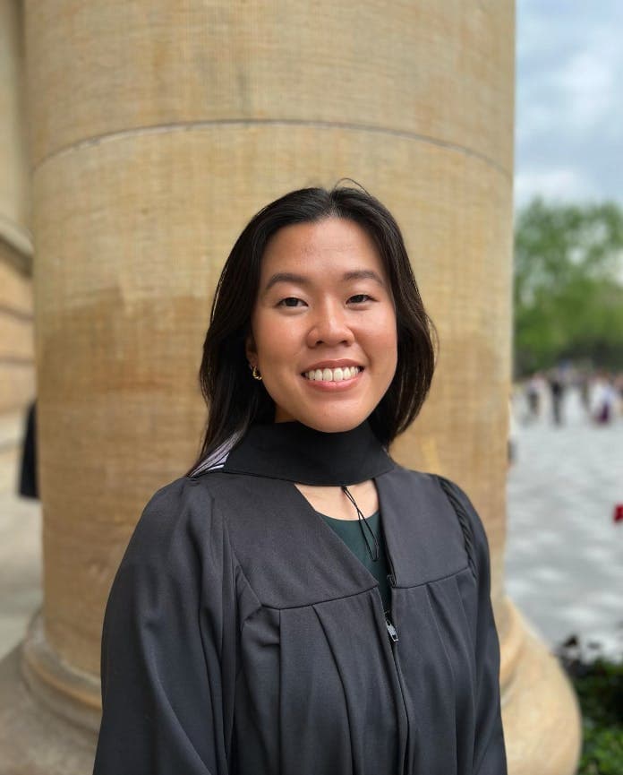 A person in a graduation gown smiles, standing in front of a pillar with a partially blurred outdoor setting in the background.