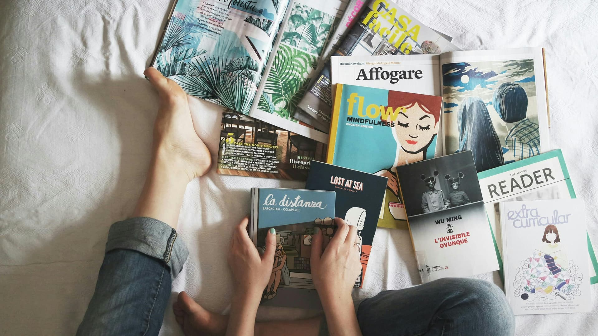 A person sitting on a bed with crossed legs is surrounded by several open books and magazines. Their hands are holding a book titled "La Distanza." The titles of the other visible publications include "Affogare," "Flow Mindfulness," "Lost at Sea," "The Happy Reader," and "Extra Curricular." The scene has a casual, relaxed vibe with a mix of art and photography in the publications.