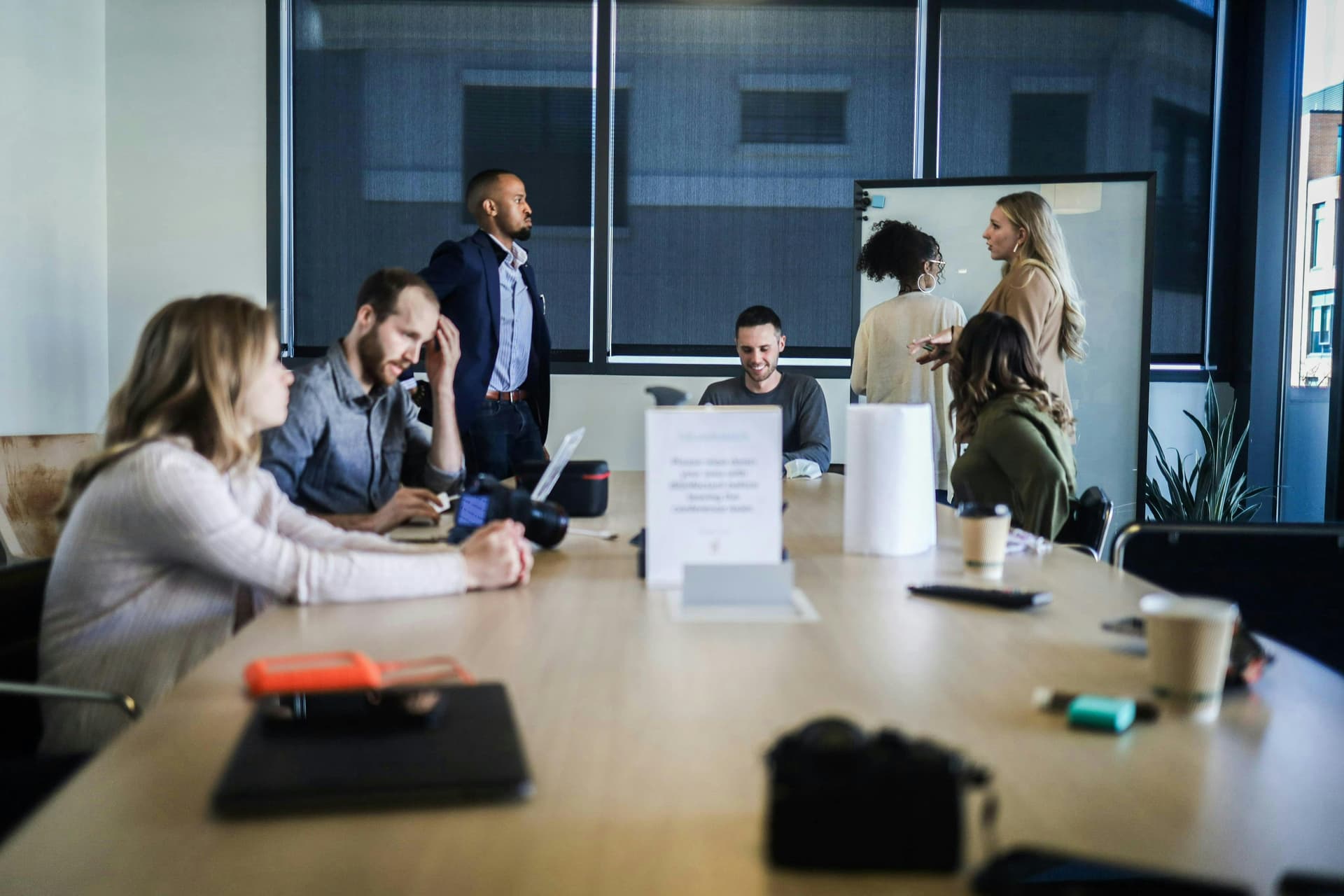 A group of people in a modern meeting room, some seated and working at a large table with laptops and cameras, while others stand and converse. The atmosphere is casual, with natural light filtering through large windows in the background.