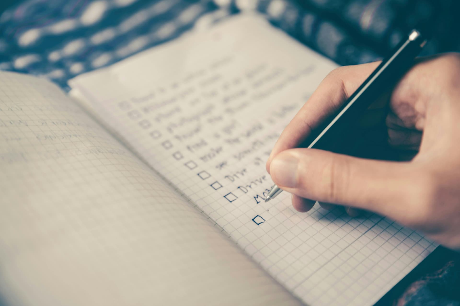 A person is writing in a notebook with a checklist of tasks, using a pen to mark off completed items. The notebook has a grid pattern, and the handwriting is slightly blurred.