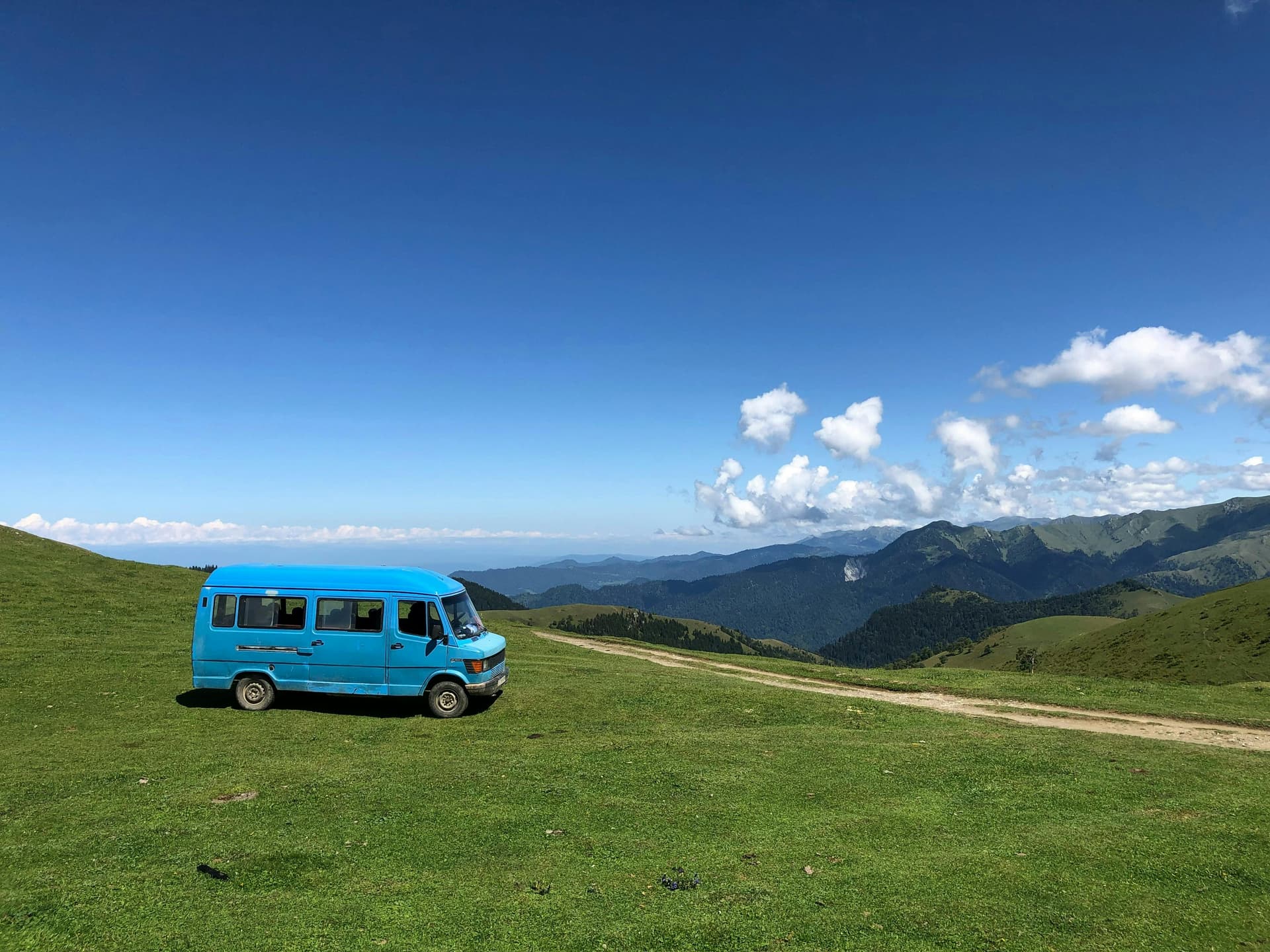 A bright blue van parked on a lush green hillside under a clear blue sky, with sweeping views of distant mountain ranges and scattered white clouds
